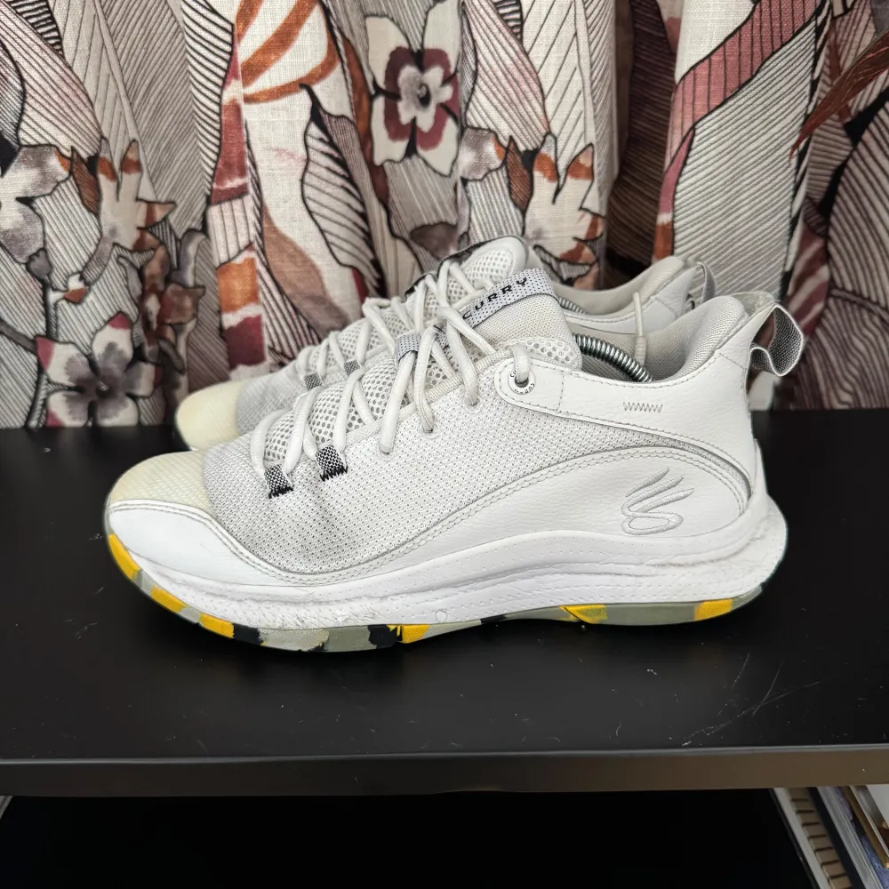 Snygga vita sneakers från Under armour med snörning och en unik sula i gult, svart och grått mönster. Perfekta för en stilren look med en twist. Skorna har en modern design med en diskret logga på sidan.. Kengät.