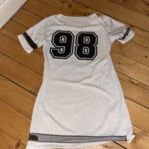 Vit klänning med nummertryck - Säljer en cool vit klänning med stort nummertryck '98' på framsidan. Klänningen har korta ärmar med svarta ränder och en avslappnad passform. Perfekt för en sportig look!
