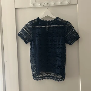 Marinblå spetsblus - Snygg marinblå spetsblus med korta ärmar och ett vackert mönster. Perfekt för att lägga till en elegant touch till din outfit. Blusen har en knappdetalj i nacken och är lätt att matcha med olika stilar.