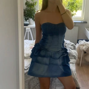 Blå axelbandslös klänning i jeans - Snygg axelbandslös klänning i blått jeansmaterial med volangdetaljer. Perfekt för en somrig look. Klänningen har en kort längd och en tight passform som framhäver figuren.