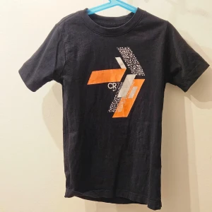 Svart t-shirt från Nike (CR7) - Säljer en svart t-shirt från Nike med ett coolt grafiskt tryck i orange och vitt på framsidan. T-shirten är kortärmad och perfekt för en sportig look.