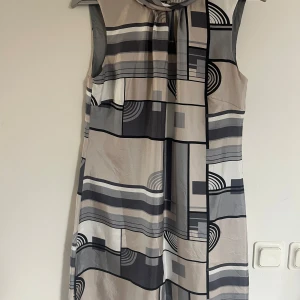 Mönstrad ärmlös klänning från Oasis - Snygg ärmlös klänning från Oasis med geometriskt mönster i grå, svart och beige. Perfekt för en stilren look. Klänningen har en knytning vid halsen som ger en extra detalj i ryggen. Materialet är 100% siden.