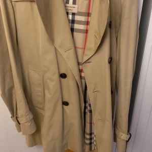 Beige trenchcoat från Burberry - Säljer en klassisk beige trenchcoat från Burberry, modell The Kensington. Jackan har ett stilrent rutigt foder och knappar framtill. Den är utrustad med ett bälte i midjan och har långa ärmar med justerbara remmar vid ärmsluten.