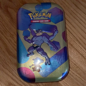 Pokémon Trading Card Game Tin 151 made in USA - Säljer en Pokémon Trading Card Game plåtlåda med Machamp-design. Perfekt för att förvara dina kort eller som samlarobjekt. Lådan har en färgglad design med hexagonmönster och är en del av Scarlet & Violet-serien.