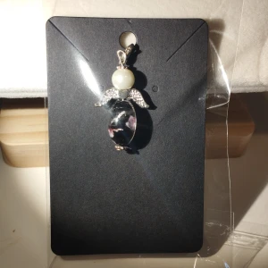 Lyckoängel hängsmycke med pärla - Unikt ängelformat hängsmycke med en vit pärla som huvud och silverfärgade vingar. Kroppen är en svart och lila mönstrad pärla. Perfekt för att ge en personlig touch till ditt halsband eller armband. Cirka 5 cm stor.