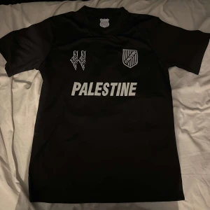 Svart fotbollströja från FC Palestina - Säljer en svart fotbollströja från FC Palestina med vit text och emblem på framsidan. Tröjan har korta ärmar och en stilren design. Perfekt för fotbollsfans som vill visa sitt stöd.