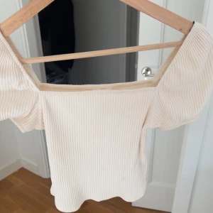 Beige ribbad offshoulder topp - Säljer en snygg beige ribbad offshoulder topp med korta puffärmar. Perfekt för en stilren look. Passar bra till både jeans och kjol.