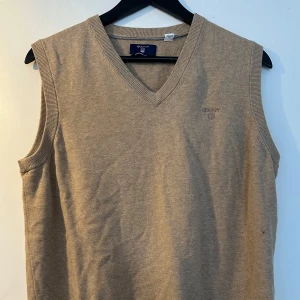 Beige stickad väst från GANT - Säljer en stilren beige stickad väst från GANT i premium bomull. Västen har en v-ringad design och ribbade kanter. Perfekt för lager-på-lager-stil.