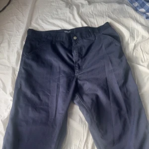 Mörkblå byxor från Carhartt WIP - Snygga mörkblå byxor från Carhartt WIP med enkel design. Byxorna har en dragkedja och knapp framtill. Perfekta för en stilren look. Nypris är cirka 1000kr