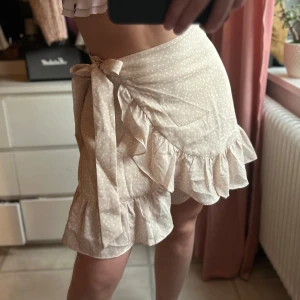 Prickig omlottkjol med volang X2 - Snygg prickig omlottkjol med volangdetaljer. Finns i både beige och svart med vita prickar. Kjolen har en knytning i sidan och är perfekt för en somrig look. Den korta längden och volangerna ger en lekfull stil.