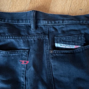 Svarta jeans från Diesel - Snygga svarta jeans från Diesel med klassisk design och röd detalj vid fickan. Perfekta för en stilren look. De har en normal passform och är tillverkade i ett slitstarkt material. Storleken säger 33 men passar mig som vanligtvis har 32/32. Därav är längden ungefär 32