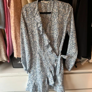 Blå blommig omlottklänning från ONLY - Säljer en söt blå omlottklänning från ONLY med vita blommor. Klänningen har långa ärmar och volangdetaljer längs kanten.