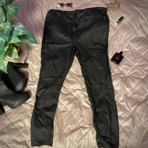 Svarta skinbyxor med knappdetalj - Snygga svarta byxor med en stilren knappdetalj i midjan. Byxorna har en klassisk design med fickor och är perfekta för en chic look. Materialet ser ut att vara syntetiskt vilket ger en glansig finish.Inte äkta skin!  Använd bara några gånger! Mer av en XXL Öppen för prisförslag 