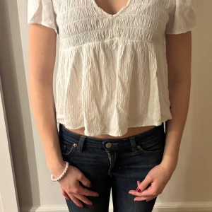 Vit blus från Brownie - Säljer en söt vit blus från Brownie med korta ärmar och v-ringning. Blusen har en luftig passform och smockdetaljer upptill som ger en fin struktur. Perfekt för en avslappnad stil.