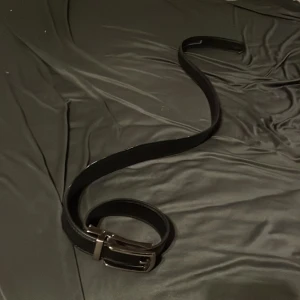 Svart bälte med silverfärgat spänne - Snyggt svart bälte med ett stilrent silverfärgat spänne. Perfekt för att ge en elegant touch till din outfit. Tillverkat i ett material som ser ut som skinn, vilket ger en klassisk look.