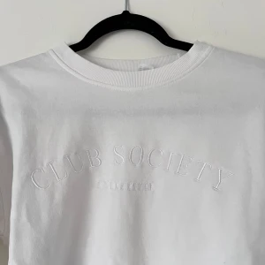 Vit sweatshirt - Säljer en stilren vit sweatshirt från Club Society med broderad text på framsidan. Tröjan har långa ärmar och ribbade muddar vid ärmslut och nederkant. Perfekt för en avslappnad look. Finns ett litet märke på baksidan av vänster axel! Önskar du veta exakta mått? Tveka inte att höra av dig!