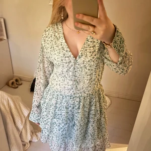 Blommig klänning med volangdetaljer - Säljer en söt blommig klänning i ljusgrön och vit med v-ringning och volangdetaljer. Klänningen har långa ärmar och en luftig passform, perfekt för vår och sommar. Nypris 599