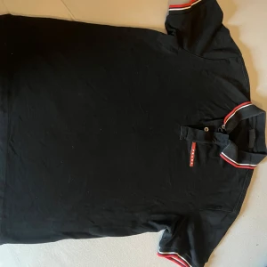 Svart pikétröja från Prada - Snygg svart pikétröja från Prada med röda och vita detaljer på krage och ärmslut. Tröjan har en klassisk pikédesign med knappar vid halsen och en diskret logga på bröstet. Perfekt för en stilren look.