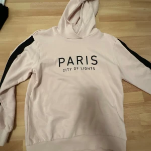 Beige hoodie med text - Säljer en ljusrosa hoodie med texten 'PARIS CITY OF LIGHTS' på framsidan. Tröjan har svarta detaljer längs ärmarna och en bekväm huva. Perfekt för en avslappnad stil.