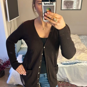 Brun kofta från Smichi Collection - Säljer en brun kofta från Smichi Collection. Den har en v-ringad design med knappar framtill. Koftan är lång i storlek L.