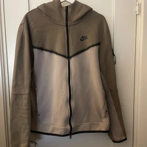 Nike tech fleece  - Säljer en snygg beige och brun huvtröja från Nike med dragkedja framtill. Tröjan har en huva och långa ärmar, perfekt för en avslappnad stil. Den har en diskret Nike-logga på bröstet.