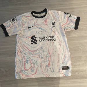 Vit fotbollströja från Nike - Säljer en vit fotbollströja från Nike med ett abstrakt mönster i rött och blått. Tröjan har korta ärmar med svarta muddar och tryck på både fram- och baksidan. Perfekt för fotbollsfans! ⚽