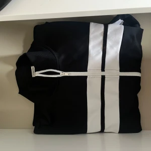 Svart och vit tröja med dragkedja - Snygg svart tröja med vita ränder och dragkedja framtill. Perfekt för en sportig look. Jackan har en hög krage och är designad för att ge en modern stil.