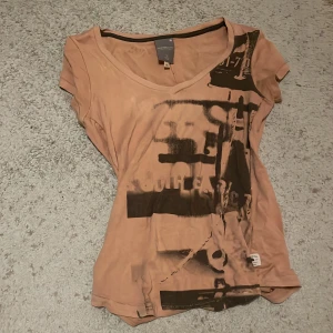 Beige y2k t-shirt med tryck - Säljer en beige t-shirt med ett unikt tryck i svart. T-shirten har en v-ringad hals och korta ärmar. Perfekt för en y2k stil.