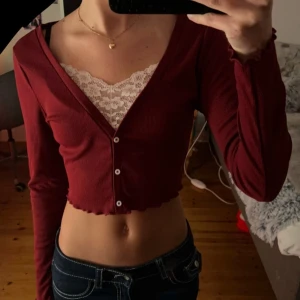 Vinröd croppad kofta med spetsdetalj - Säljer en snygg vinröd croppad topp/tröja med spetsdetalj vid halsen. Tröjan har långa ärmar och knappar framtill. Perfekt för en varm sommardag