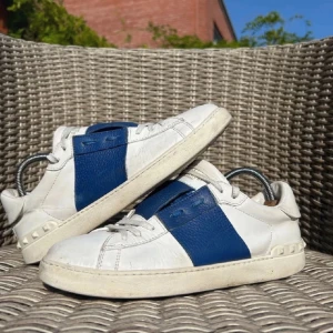 Vit och blå sneaker - Snygga sneakers i vitt med en bred blå detalj på sidan. Skorna har en klassisk design med snörning och en bekväm sula. Perfekta för en stilren look.
