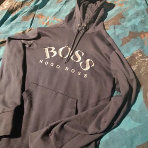 Mörkblå hoodie från Hugo Boss - Säljer en snygg mörkblå hoodie från Hugo Boss med stor logga på bröstet. Tröjan har en praktisk dragkedja vid ärmslutet och en klassisk känguruficka framtill. Perfekt för en stilren och bekväm look. Storlek M. Pris kan diskuteras inga hål eller fläckar.