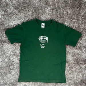 Grön t-shirt  Nike x Stussy - Säljer en grön t-shirt från Nike x Stussy med tryck av olika städer på framsidan. T-shirten har en klassisk rund hals och korta ärmar. Perfekt för en avslappnad stil. Storlek S/M
