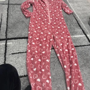 Rosa fleece onesie med hjärtan - Supermysig rosa fleece onesie med vita hjärtan och små korsmönster. Dragkedja framtill gör den enkel att ta på och av. Perfekt för chill hemma eller sleepover med kompisar. Långärmad och helkroppsdesign för extra komfort.