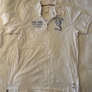 Vit Esprit t-shirt med tryck och krage - Vit t-shirt från Esprit med klassisk pikékrage och knappslå. Tryck på bröstet med texten 'San Diego Open Air Tour Calif.' och 'Festival Vibe No.3'. Små detaljer i blått och svart, samt en etikett vid nederkanten. Perfekt för dig som gillar festivalstil.