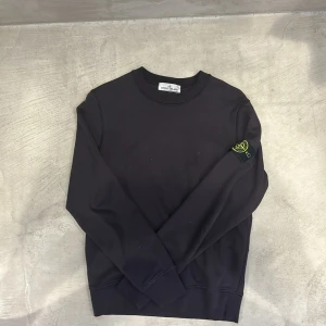 Svart/lila  sweatshirt från Stone Island - Svart/lila crewneck sweatshirt från Stone Island med klassisk rund hals och ikonisk logotyp-patch på vänster ärm. Tröjan har ribbade muddar och är tillverkad i mjuk bomull som är skön att bära. Perfekt för dig som gillar stilrena och snygga streetwear-plagg.