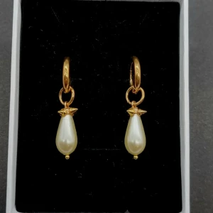 Guldörhängen med droppformad pärla - Snygga örhängen i guldton med droppformade vita pärlor som hänger från små ringar. Detaljerad spetsdesign ovanför pärlan ger en cool och elegant vibe. Perfekta för att lyfta din stil med en touch av lyx och trend.