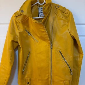 Gul skinnjacka från Zara Girls - Cool gul skinnjacka från Zara Girls med silvriga dragkedjor och detaljer. Jackan har klassisk bikerstil med sned dragkedja, axelklaffar och tryckknappar. Insidan har tryck med texten 'ROCK'. Perfekt för dig som vill sticka ut med färg och attityd.