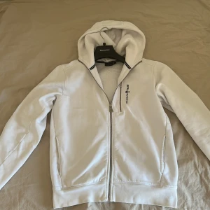 Vit hoodie från Sale Racing med dragkedja - Vit hoodie från Same Racing med hel dragkedja framtill och huva. Tröjan har långa ärmar, två sidofickor och en stilren logga på bröstet. Perfekt för chill dagar eller när du vill ha en clean look. Materialet känns mjukt och bekvämt mot huden.