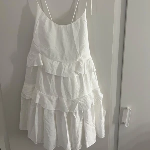 Vit volangklänning från Nelly - Supersöt vit kortklänning från Nelly med flera lager volanger och tunna axelband. Klänningen har en öppen rygg och är tillverkad i ett lätt material som ger en luftig känsla. Perfekt för dig som gillar romantisk och trendig stil.