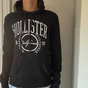 Svart hoodie från Hollister - Svart hoodie från Hollister med stor vit logga och tryck på bröstet. Inga defekter eller skador. Modellen är runt 175 cm. Pris ej hugget i sten. Perfekt för chill dagar och har en klassisk streetstil.