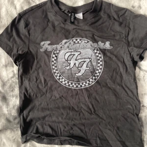 Svart Foo Fighters t-shirt Divided XXS - Svart crop t-shirt från H&M Divided i storlek XXS med Foo Fighters-tryck i silver på bröstet. Rund hals och korta ärmar, tillverkad i mjuk bomull. Snyggt grafiskt tryck med bandets logga och schackrutigt mönster runt om.