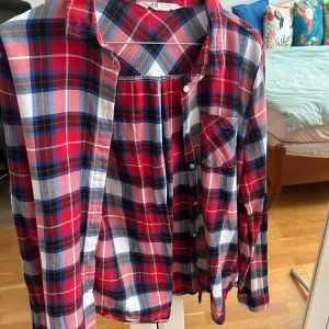 Rutig röd skjorta från H&M - Säljer en klassisk rutig skjorta från H&M i rött, blått och vitt. Skjortan har lång ärm, knappar framtill och en bröstficka. Perfekt för dig som gillar avslappnad stil med lite edge. Materialet är mjuk bomull som känns skönt mot huden.