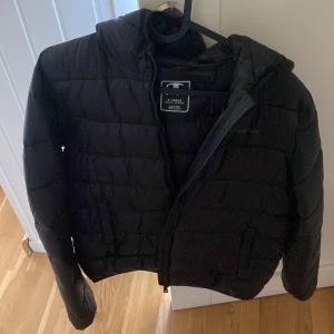 Svart pufferjacka från Champion XL - Svart pufferjacka från Champion i storlek XL (162/167). Jackan har huva, quiltad design och dragkedja framtill. Perfekt för kalla dagar när du vill hålla stilen och ändå vara varm. Enkel och clean look som funkar till det mesta.