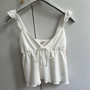 Vit frilltop från Nelly med knyt - Helt ny. Supersöt vit frilltop från Nelly i storlek XS. Toppen har volangkant, v-ringning och knyt framtill. 