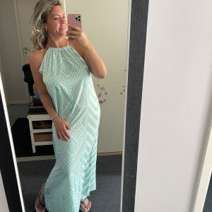 Mintgrön långklänning med mönster - Säljer en mintgrön långklänning med vitt geometriskt mönster. Klänningen har halterneck och är ärmlös, med en loose passform och slits på sidan. Perfekt för varma dagar och chill vibes. Materialet känns lätt och luftigt.helt ny