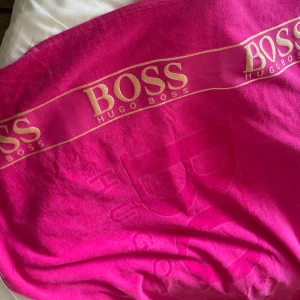  Hugo Boss  - Stor och lyxig strandhandduk från Hugo Boss i en riktigt poppig rosa färg. Handduken har tydlig logga i guld och präglad text över hela ytan. Perfekt för stranden eller poolen när du vill sticka ut och visa stil.