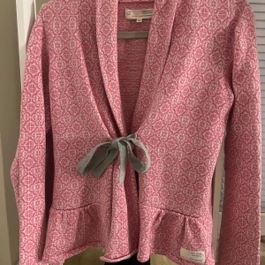 Rosa cardigan jacka Odd Molly - Superfin rosa och vit cardigan jacka från Odd Molly med snyggt mönster och grå knytband framtill. Jackan har långa ärmar, två fickor och volangdetalj nedtill.