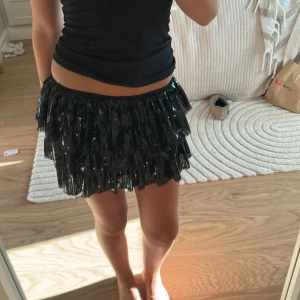 Svart paljettkjol med volanger - Supersnygg svart minikjol med glittrande paljetter och flera lager volanger. Kjolen har en tight passform i midjan och ger en riktigt festlig vibe med sitt skimrande material. Perfekt för dig som vill sticka ut och älskar att glittra.