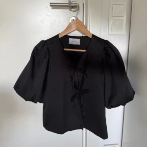 Svart blus med puffärm från Neo Noir - Supersnygg svart blus från Neo Noir med puffiga korta ärmar och knytning framtill. Blusen har en lätt glansig yta. Storlek 34 men passar även 36. Nypris 599 kr