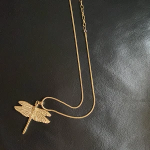 Guldigt trollsländehalsband från IAKOU  - Snyggt halsband i guld med en detaljrik trollslända som hänge. Kedjan är tunn och elegant, perfekt för att ge din outfit en cool och unik touch. Passar dig som gillar naturinspirerade smycken och vill sticka ut lite extra.